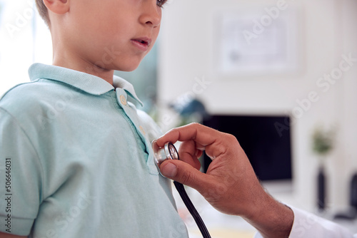 Close Up Of Male Doctor Or GP Wearing White Coat Examining Boy Listening To Chest With Stethoscope