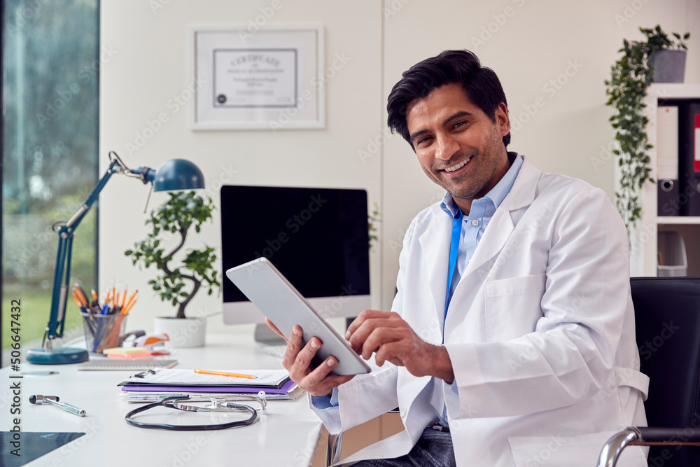 Portrait Of Male Doctor Or GP Wearing White Coat Sitting At Desk In Office Using Digital Tablet