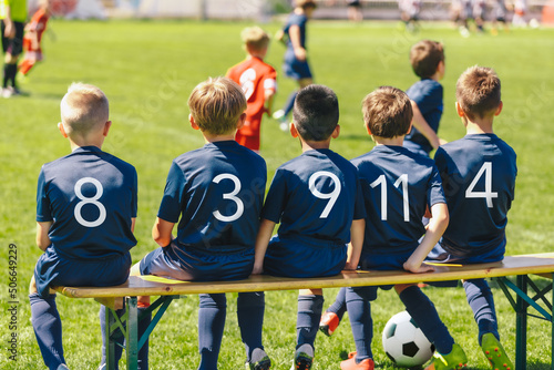 Football players wearing blue jersey shirts in the youth team sitting on a wooden bench. Multiracial group of kids in a school sports team. Boys in blue jersey shirts watching a tournament game