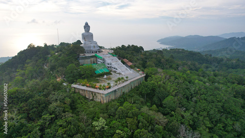 Drone view of the Big Buddha  Thailand. The Big Buddha is sitting on hill in the lotus position  meditating. The sun shines brightly through clouds. There s a jungle all around. A ray of sun on water