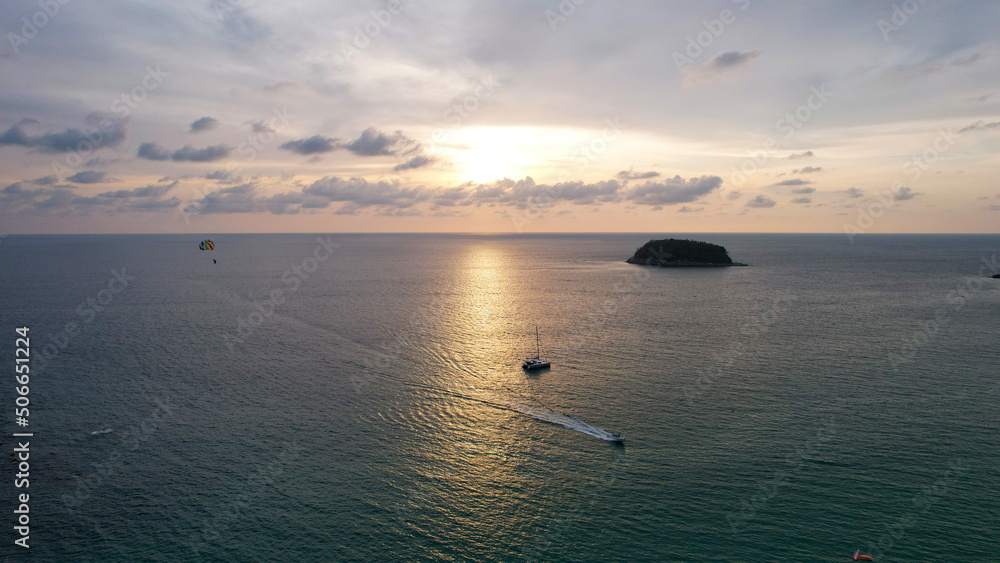 Parasailing at sunset with a view of the island. Parachute flight in the midst of a colorful sunset. Small waves, people relax on the beach, palm trees grow, there are hotels. Boats float. Asia Phuket