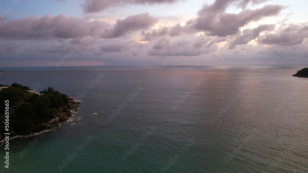 View from the top of Phuket island and the sea. It's raining in the distance, grey clouds. In places, you can see the blue sky and sunset. There are houses and green trees on island. Boats are sailing