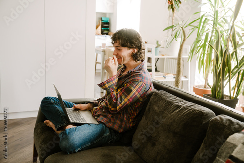 White man wearing shirt using laptop while resting on couch © Drobot Dean