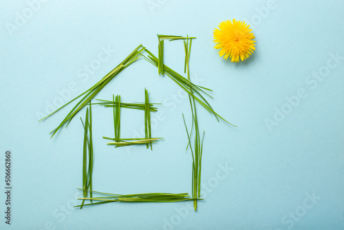 house made of grass bades with chimney and a dandelion as the sun. Grass straws on light blue background photo