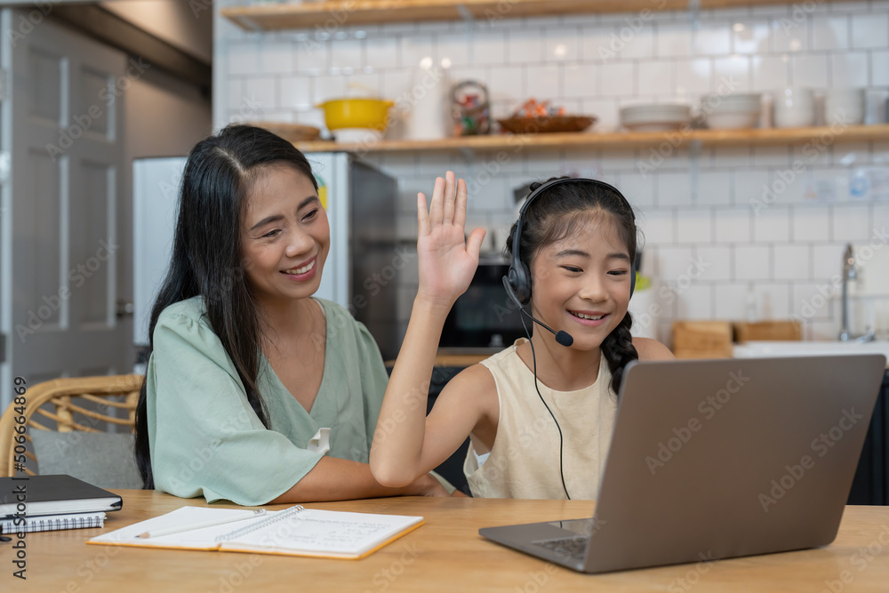 Happy asian mother and child sitting at kitchen table with colored pencils, attending virtual drawing class via video call, smiling and waving hello at laptop computer screen to greet online teacher Happy asian mother and child sitting at kitchen table with colored pencils, attending virtual drawing class via video call, smiling and waving hello at laptop computer screen to greet online teacher