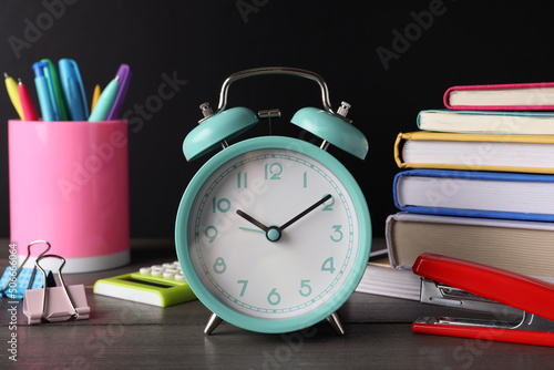 Alarm clock and different stationery on wooden table near blackboard. School time