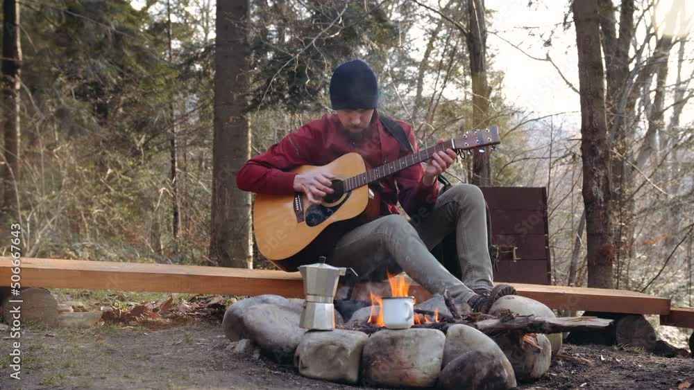 Young guy tourist in woods by the fire in a navy blue hat, playing the guitar Young guy tourist in woods by the fire in a navy blue hat, playing the guitar
