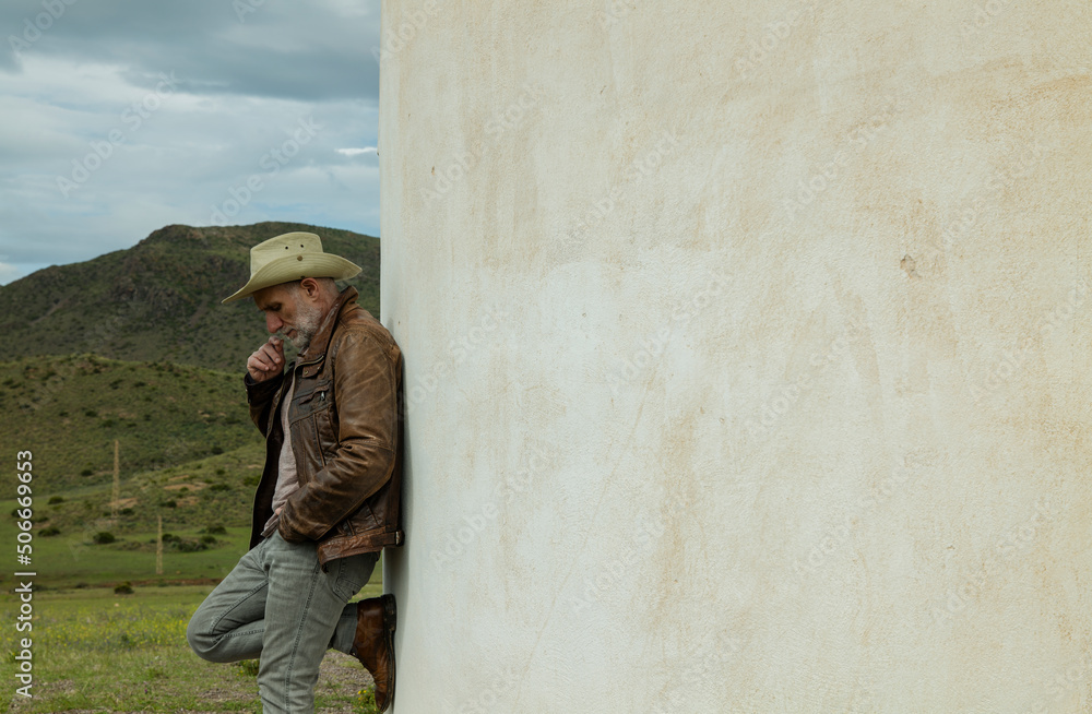 Adult man in cowboy hat standing against white wall in field Adult man in cowboy hat standing against white wall in field