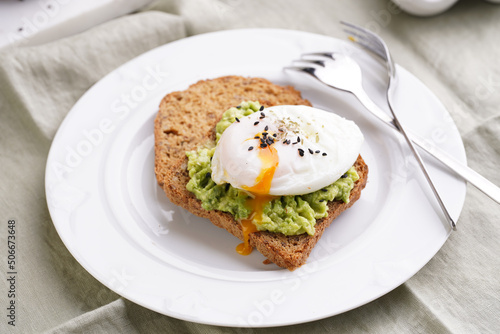 Slices of gluten-free sunflower seeds bread with mashed avocado, poached egg and sesame seeds on white plate on green checkered napkin