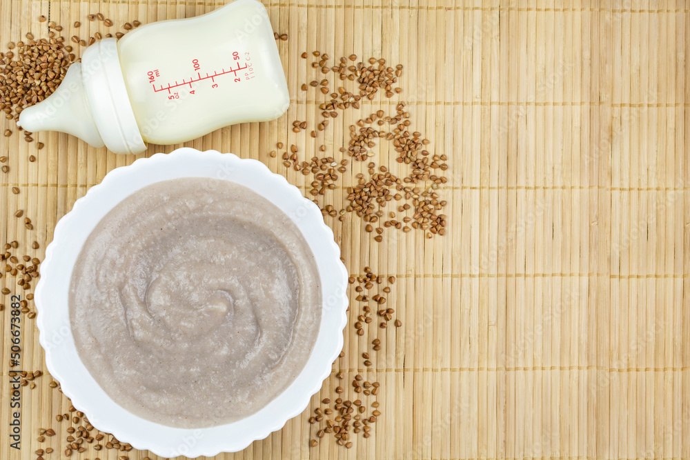 Buckwheat porridge for the baby from ground cereals in a white bowl, a bottle of milk on a brown bamboo background. Space for the text. The first complementary food of a child, baby nutrition. Buckwheat porridge for the baby from ground cereals in a white bowl, a bottle of milk on a brown bamboo background. Space for the text. The first complementary food of a child, baby nutrition.