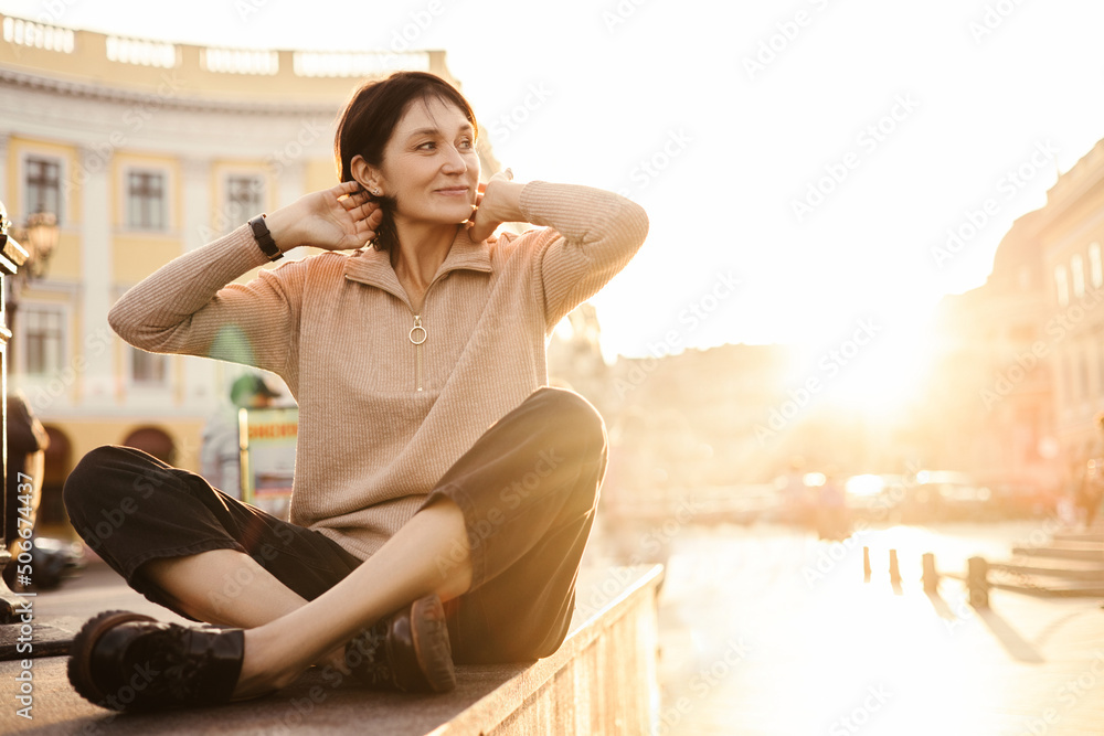 Beautiful adult caucasian woman enjoying free time at sunset outdoors. Lady with short dark haircut wears jacket and leggings. Spring lifestyle freedom concept.