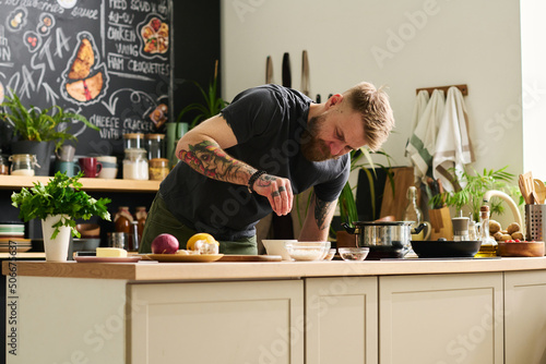 Modern young Caucasian man standing in kitchen at home cooking meal adding spices into bowl