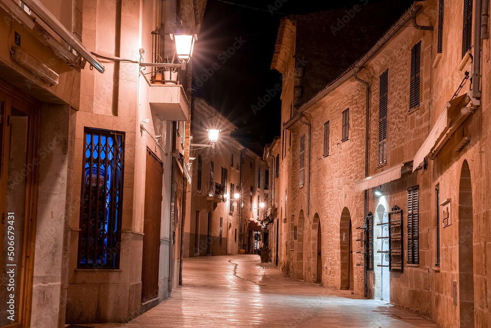 Mallorca, Spain. April 27, 2022. Illuminated alley amidst historic residential buildings. Houses and sconce lights on road in town in dark. Empty street in old city at night.
