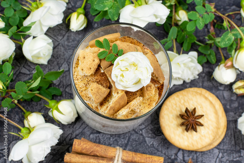 Multi-layered cottage cheese and yogurt dessert with banana, cinnamon and cookies in a glass glass and white roses on a black (dark) background. Top view.