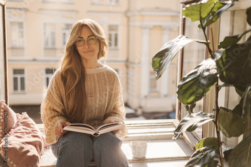 Attractive adult caucasian lady holds book in hands, looks at camera while sitting on windowsill in sunny weather. Blonde model in glasses, sweater and jeans. Leisure concept, lifestyle