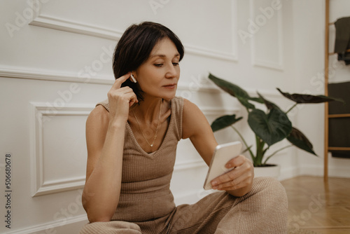 Beautiful adult caucasian woman listens to music through headphones using phone indoors. Brunette wears light brown homemade suit. Concept of spending time, hobby
