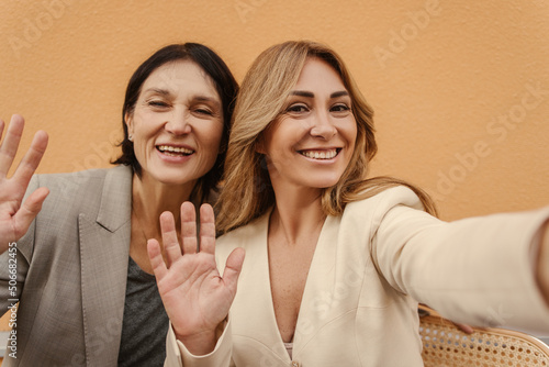 Happy caucasian adult girlfriends take photo of themselves with phone, waving their hands at camera. Pretty women in stylish clothes smile with their teeth. Enjoying little things, positive attitude