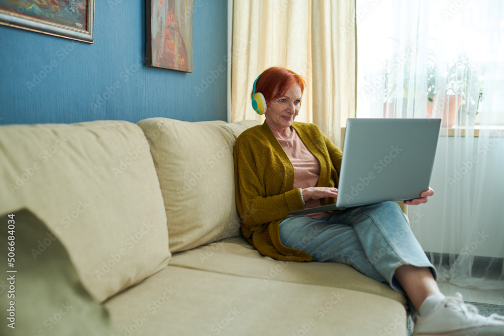 Senior woman in wireless headphones using laptop while resting on sofa in living room, she having online consultation Senior woman in wireless headphones using laptop while resting on sofa in living room, she having online consultation