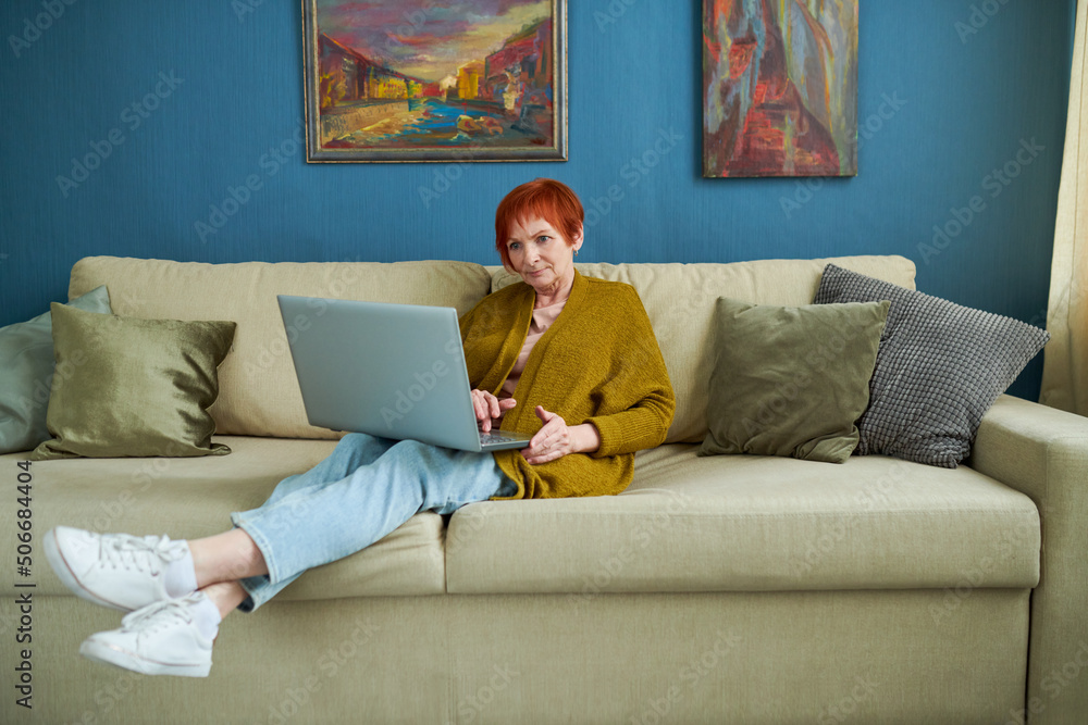 Senior woman resting on sofa in living room with laptop on her knees, she communication online with grandchildren Senior woman resting on sofa in living room with laptop on her knees, she communication online with grandchildren