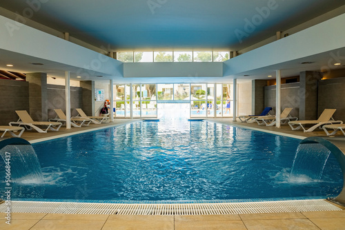 Water falling in swimming pool. Deck chairs arranged in room at luxurious resort. View of indoor blue pool in hotel during summer.