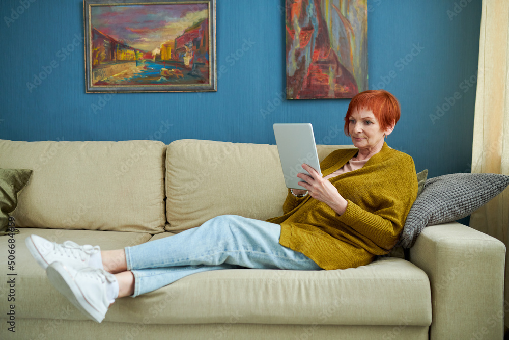 Elderly woman lying on sofa in living room and using digital tablet, she communication online or watching video