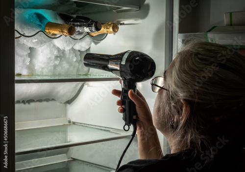 Obraz na plátne Woman defrosting the inside of a fridge where there are some bottles, with the help of a hair dryer