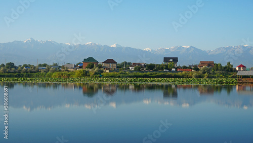 The mirror surface of the lake water reflects pink lotuses, large green water lilies, high mountains of the Trans-Ili Alatau, trees and houses. Beautiful landscape on mirror pond. Almaty, Kazakhstan