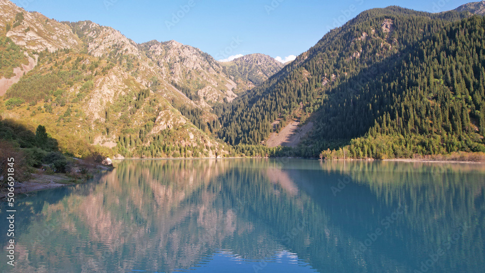 View of the mountain lake Issyk from a height. Turquoise-emerald water from the glacier. Reflection of mountains, forests and the sun on the water. The river flows into the lake. Issyk Dam. Kazakhstan