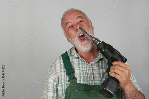 Taken literally in the nose picking. An old craftsman picks in his nose with his drilling machine, in front of a white wall. Danger! Nose picking can cause injuries.