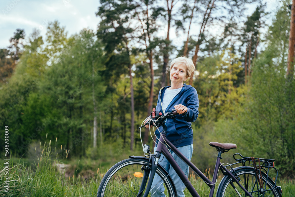Fototapeta premium Short blond hair aged woman enjoying healthy outdoor activities. Cycling. Smiling senior woman standing on a country lane with her bicycle.