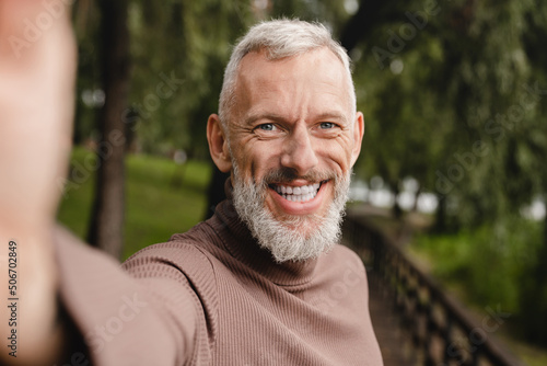 Smiling caucasian mature man looking at the camera, taking selfie on smart phone, having video call, vlogging blogging outdoors in park.