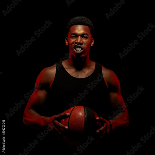 Tablou canvas Basketball player side lit with red color holding a ball against black background
