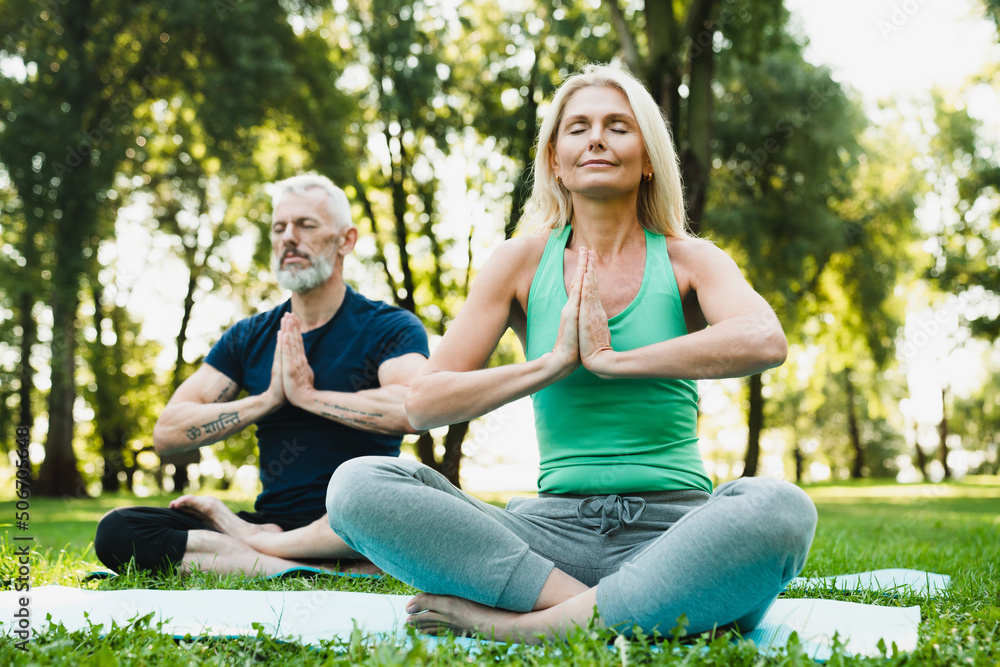 Low angle view on mature couple relaxing meditating in lotus position together feeling zen-like on fitness mat in forest park. Active seniors, healthy lifestyle. Yoga class outdoors. Low angle view on mature couple relaxing meditating in lotus position together feeling zen-like on fitness mat in forest park. Active seniors, healthy lifestyle. Yoga class outdoors.