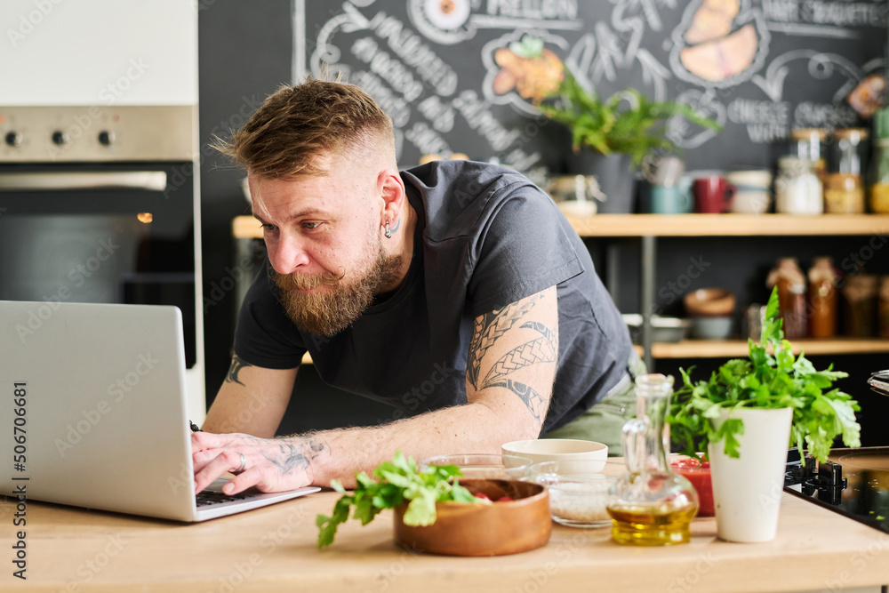 Stylish bearded Caucasian man searching meal recipe or texting message in Internet on laptop in loft kitchen at home Stylish bearded Caucasian man searching meal recipe or texting message in Internet on laptop in loft kitchen at home