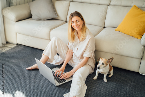 Beautiful blonde woman using laptop. working from home, studing or online shopping. Freelancer or influencer remote job. Sitting on floor in living room with small cute dog. Vertical composition © Iryna&Maya