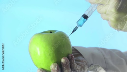 Close-up of a human in a medical coat and gloves injecting a syringe into an apple with some liquid on a blue background