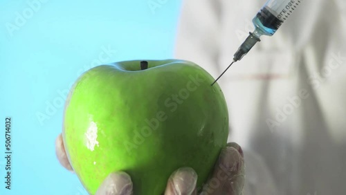 Close-up of a human in a medical coat and gloves injecting a syringe into an apple with some liquid on a blue background