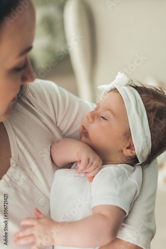 Close portrait of a little cute dark-haired newborn girl in a white suit in the arms of a young mother. Motherhood. Parent. Healthy sleep.