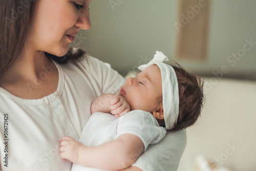 Close portrait of a little cute dark-haired newborn girl in a white suit in the arms of a young mother. Motherhood. Parent. Healthy sleep.