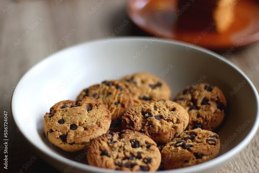 Bowl of chocolate chip cookies, plate of biscuits, open books and reading glasses on the table. Selective focus. Bowl of chocolate chip cookies, plate of biscuits, open books and reading glasses on the table. Selective focus.