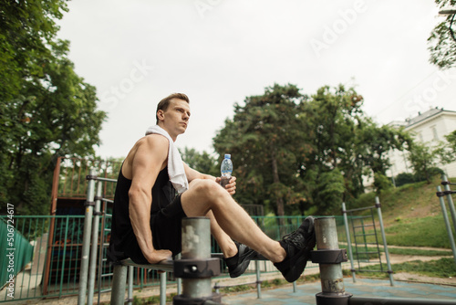 Little break. Young athletic guy with muscular body, sportsman and fitness model, strong man sitting on parallel bars. Athletic body shape, guy relax at outdoor gym.