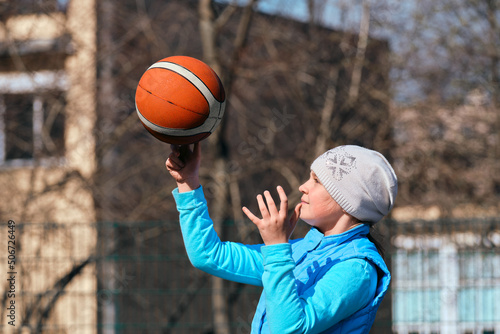 A young girl in a blue outerwear 10-11 years old holds a basketball in her hands. Child playing basketball outdoors.