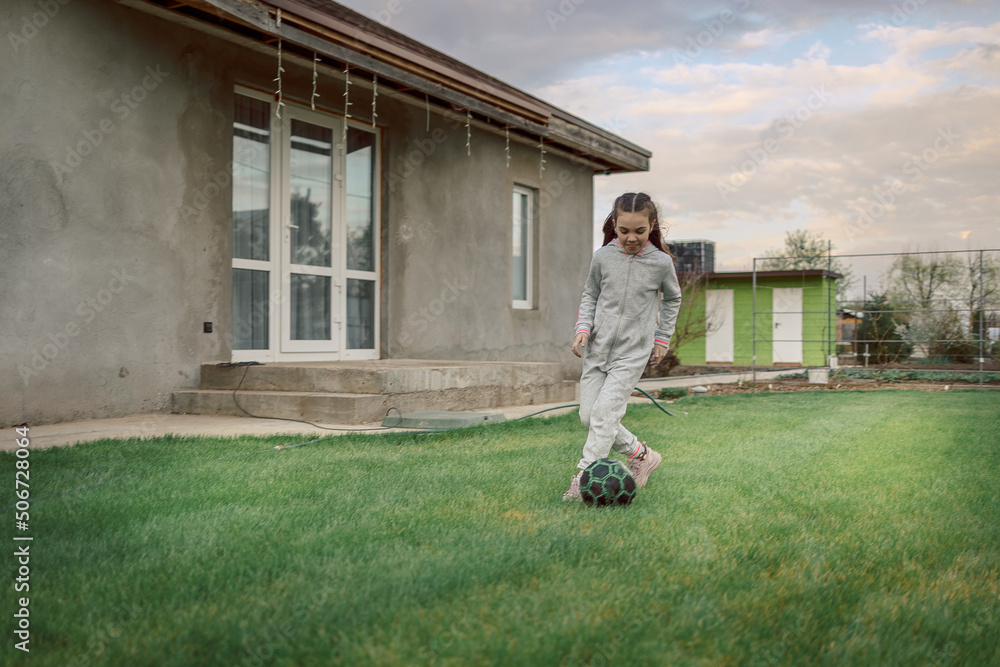 Fototapeta premium Cute little girl playing football with soccer ball on green lawn in backyard of house. Child kicking soccer ball on field. High quality photo
