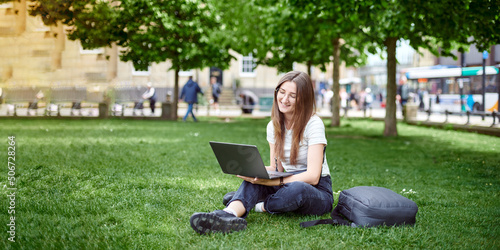 On the banner, a young girl works with a laptop in the fresh air in the park, sitting on the lawn. The concept of remote work. Work as a freelancer. The girl takes courses on a laptop and smiles. © artdolgov