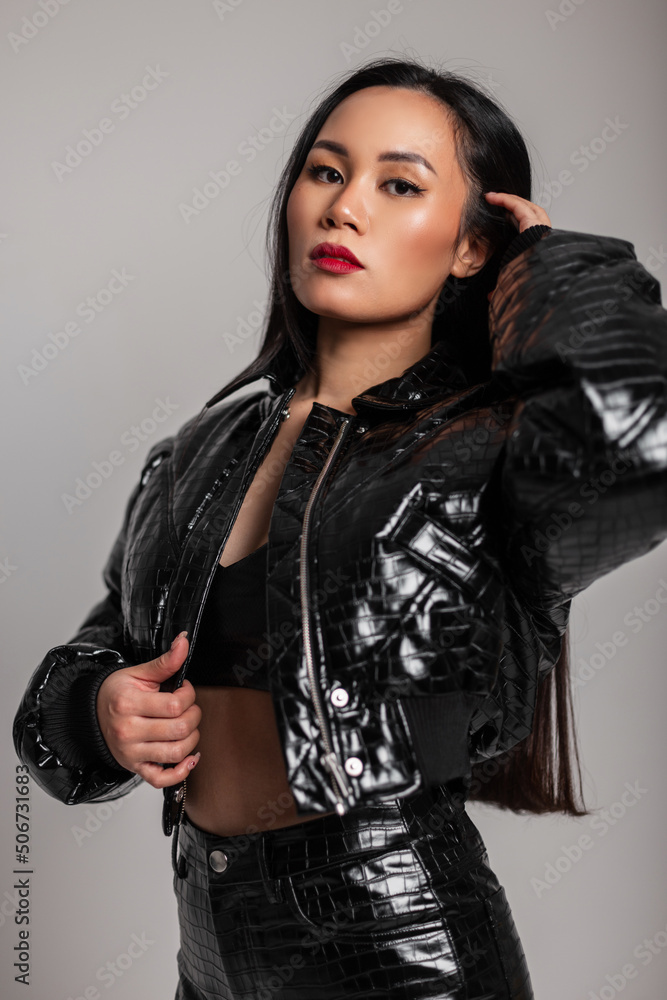 Naklejka premium Fashionable portrait of a stylish Asian girl with makeup in a fashion black leather jacket with bra and pants on a gray background