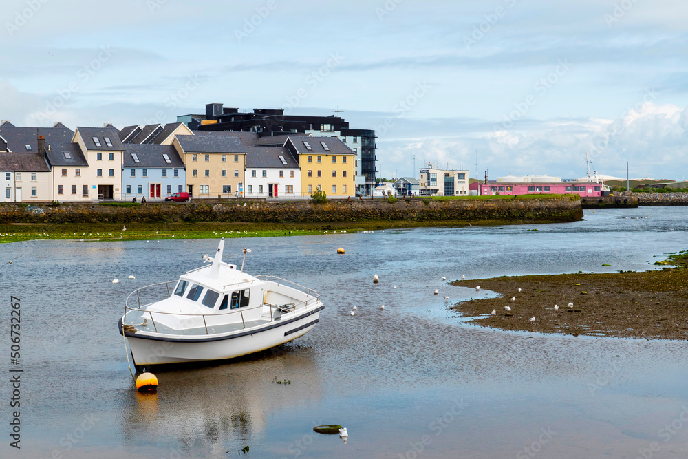 Obraz premium Small boat at low tide. River Corrib, Claddagh area in the background. Galway city Ireland. Popular town landmark and tourist attraction. Blue cloudy sky.