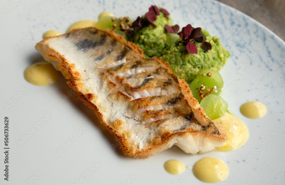 Close-up on grilled piece of fish with green puree and dark red edible plants, radish sprouts, on off white plate. Modern restaurant cuisine.