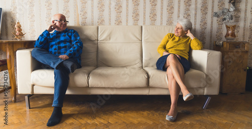 фотографія happy Caucasian grandparents sitting apart at the opposite ends of the sofa full shot indoor senior people concept