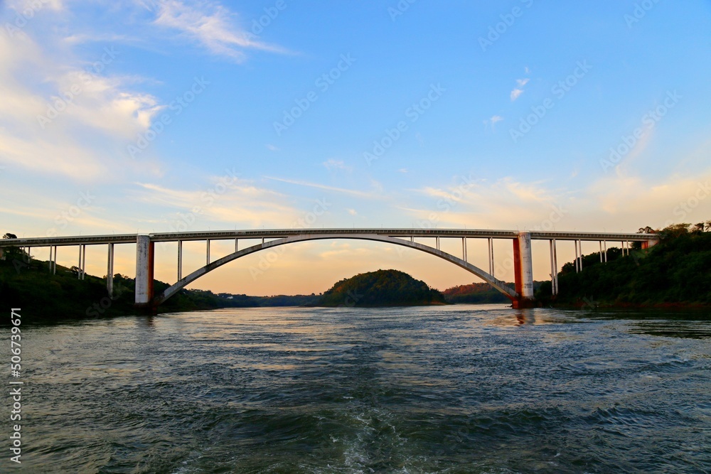Fototapeta premium Bridge over the Iguacu River on the border between Brazil and Paraguay. Arch bridge over the waters of the river and an island in the background. Dusk sky.
