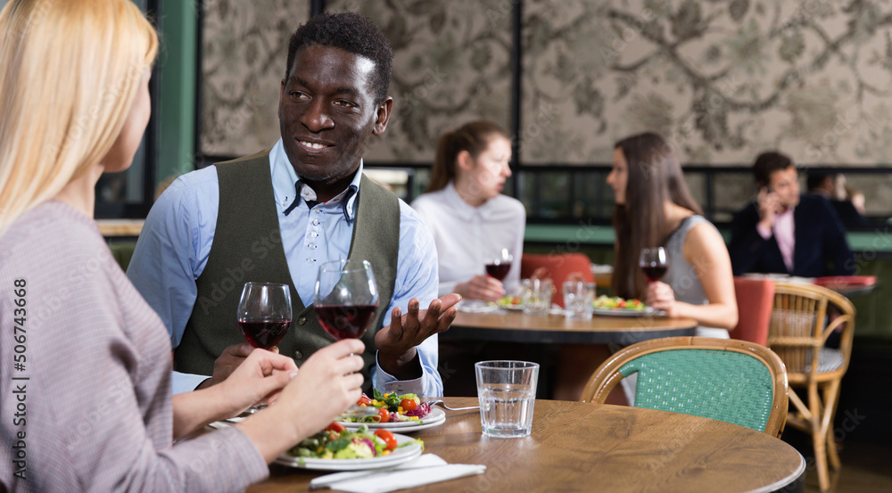 Confident African American businessman with happy cheerful positive smiling female partner having dinner and drinking red wine at restaurant